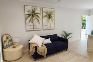 Una sala de estar con un sofá azul marino, dos cojines blancos, una manta color beige, una mesita redonda con una lámpara y dos cuadros enmarcados con imágenes de palmeras en la pared.