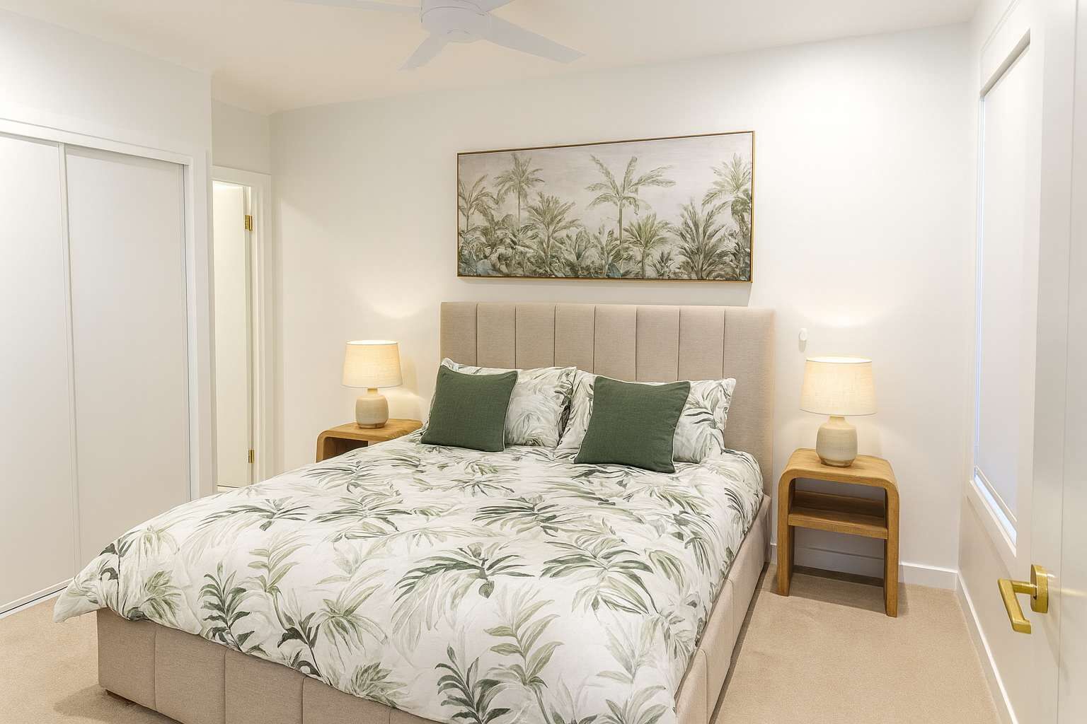 Une chambre à coucher comprenant un lit, deux tables de chevet avec des lampes, un ventilateur de plafond, une porte et un tableau accroché au mur.