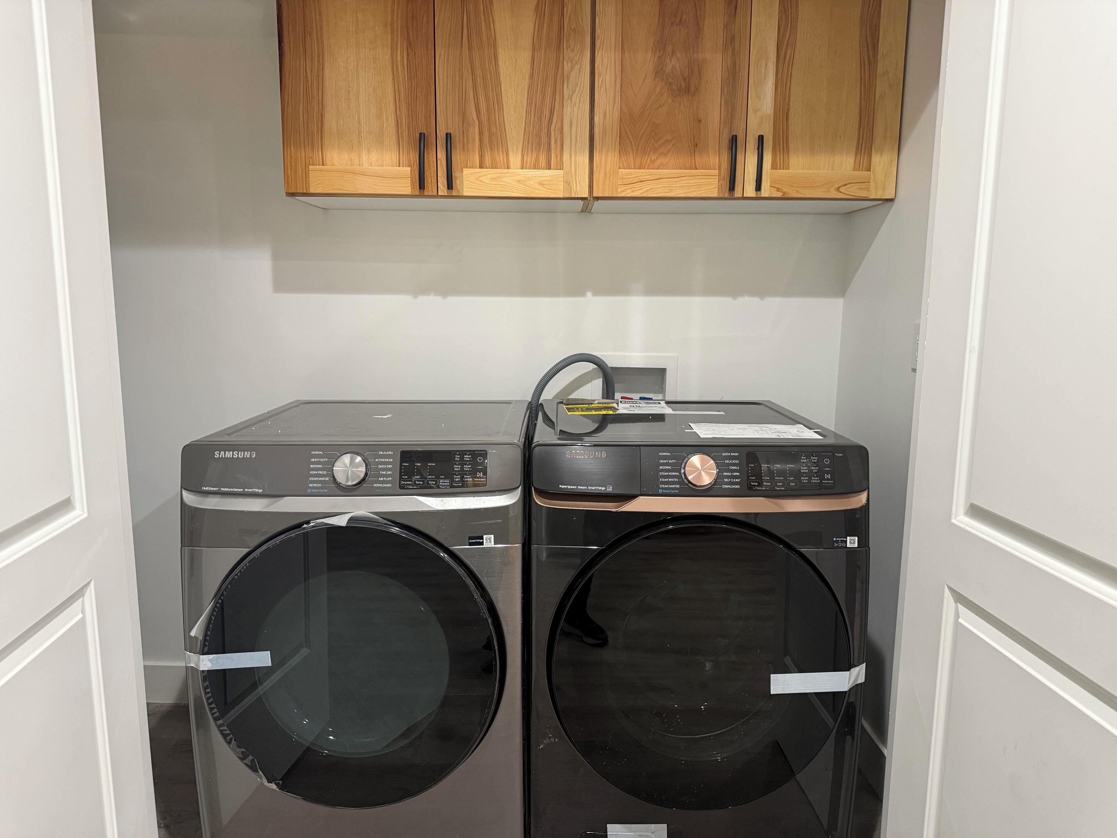 A laundry room with two front-loading washing machines and a dryer, wooden cabinets above, and a white door frame.