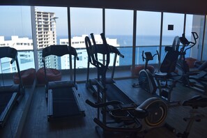 A gym with exercise equipment and a view of buildings through large windows. - Santa Marta: Sunset View, Pools, and Private Beach in Pozos Colorados (Santa Marta)