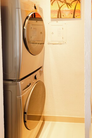 A laundry room with two washing machines, a wall-mounted electrical outlet, and a painting of a sunset with palm trees. - Santa Marta: Sunset View, Pools, and Private Beach in Pozos Colorados (Santa Marta)