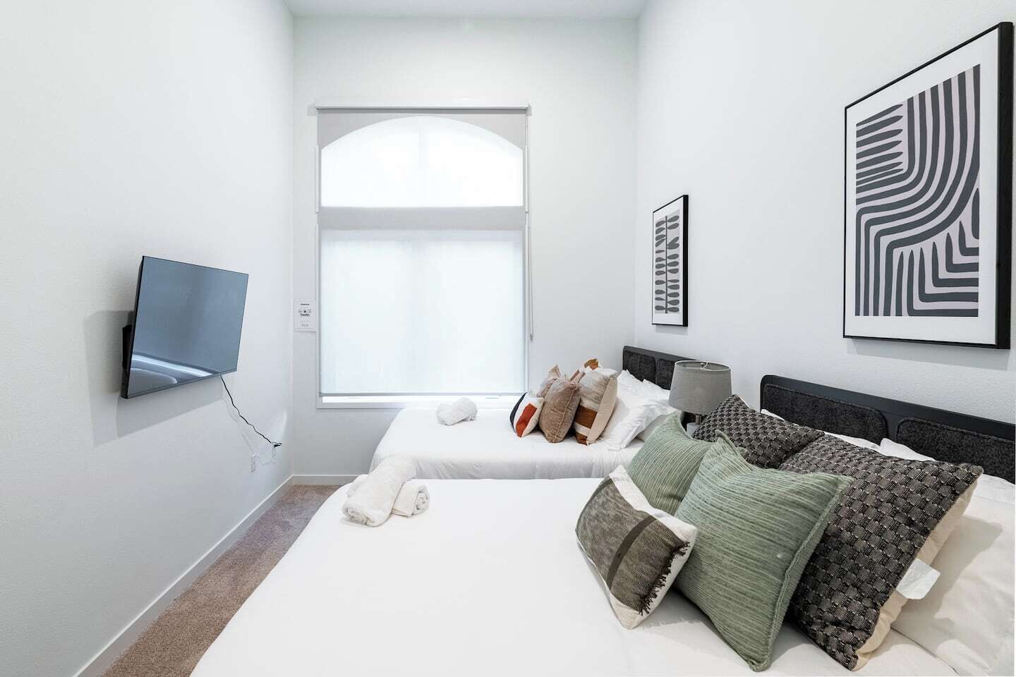 Un dormitorio moderno con un ventanal grande, un televisor de pantalla plana en la pared y dos camas con varios almohadones y una mesita de noche.