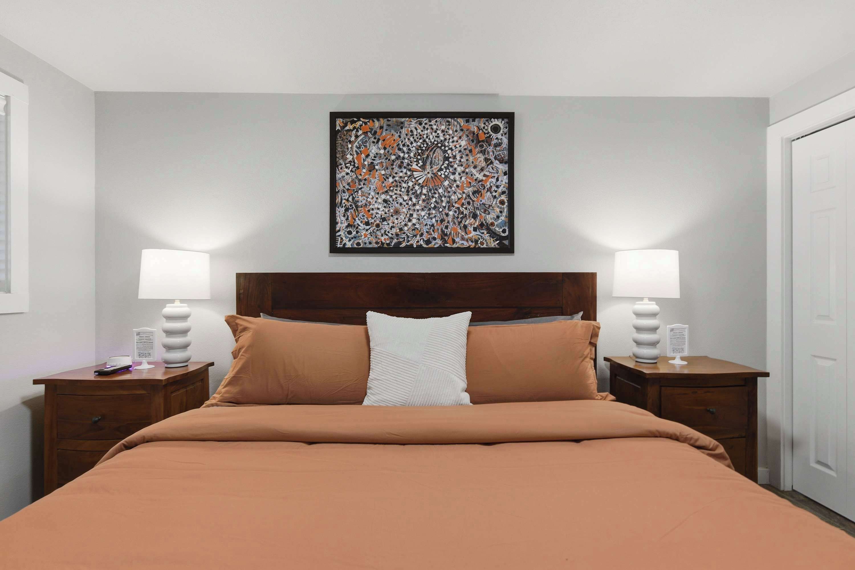 A hotel room with a bed, two bedside tables, a nightstand, a wall-mounted abstract painting, and a door.