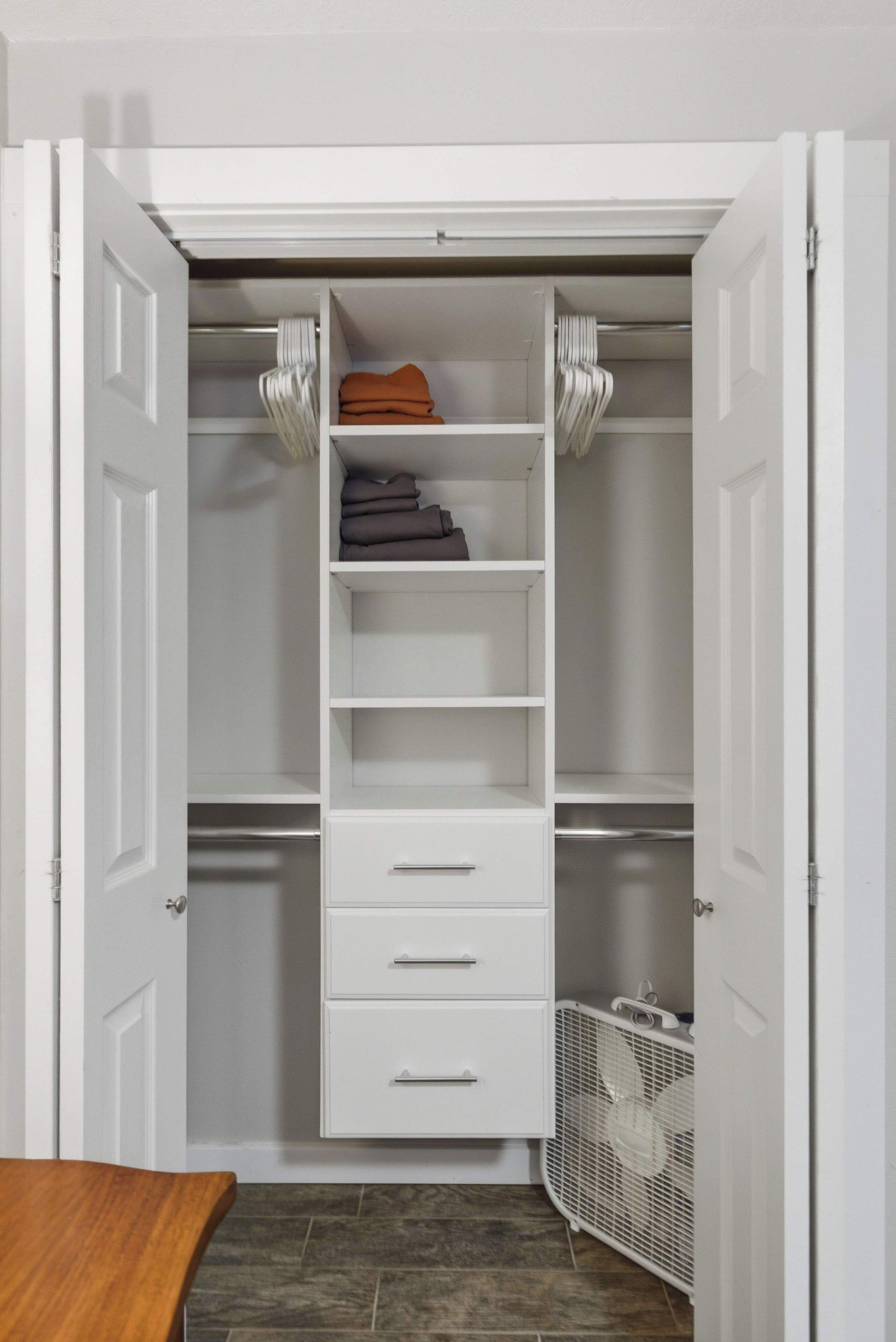 A walk-in closet with white shelving, a folding chair, and a fan.