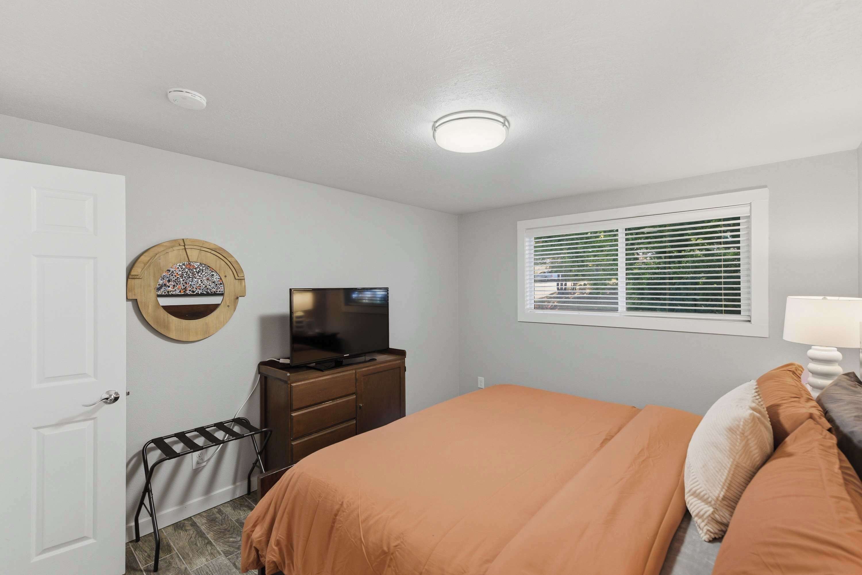 A bedroom with a bed, a television on a dresser, a window with blinds, and a door.