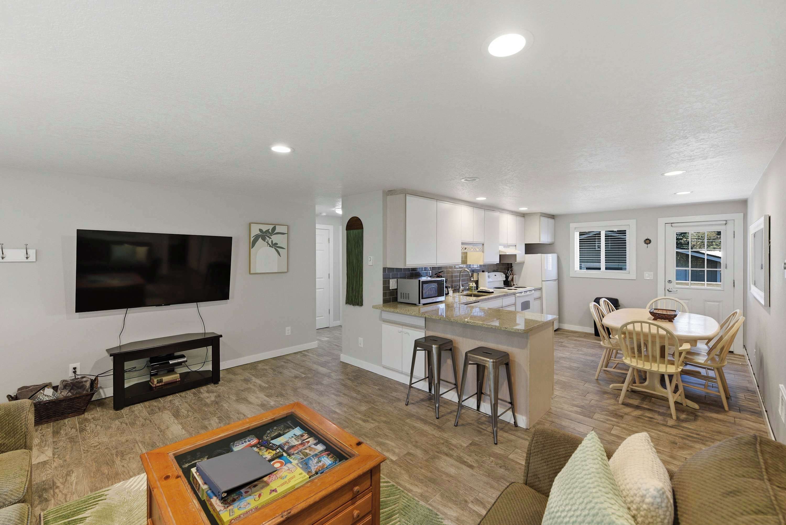 A modern living room with a flat-screen TV, a coffee table with board games, a dining area with a table and chairs, and a kitchen with white cabinets and a microwave.