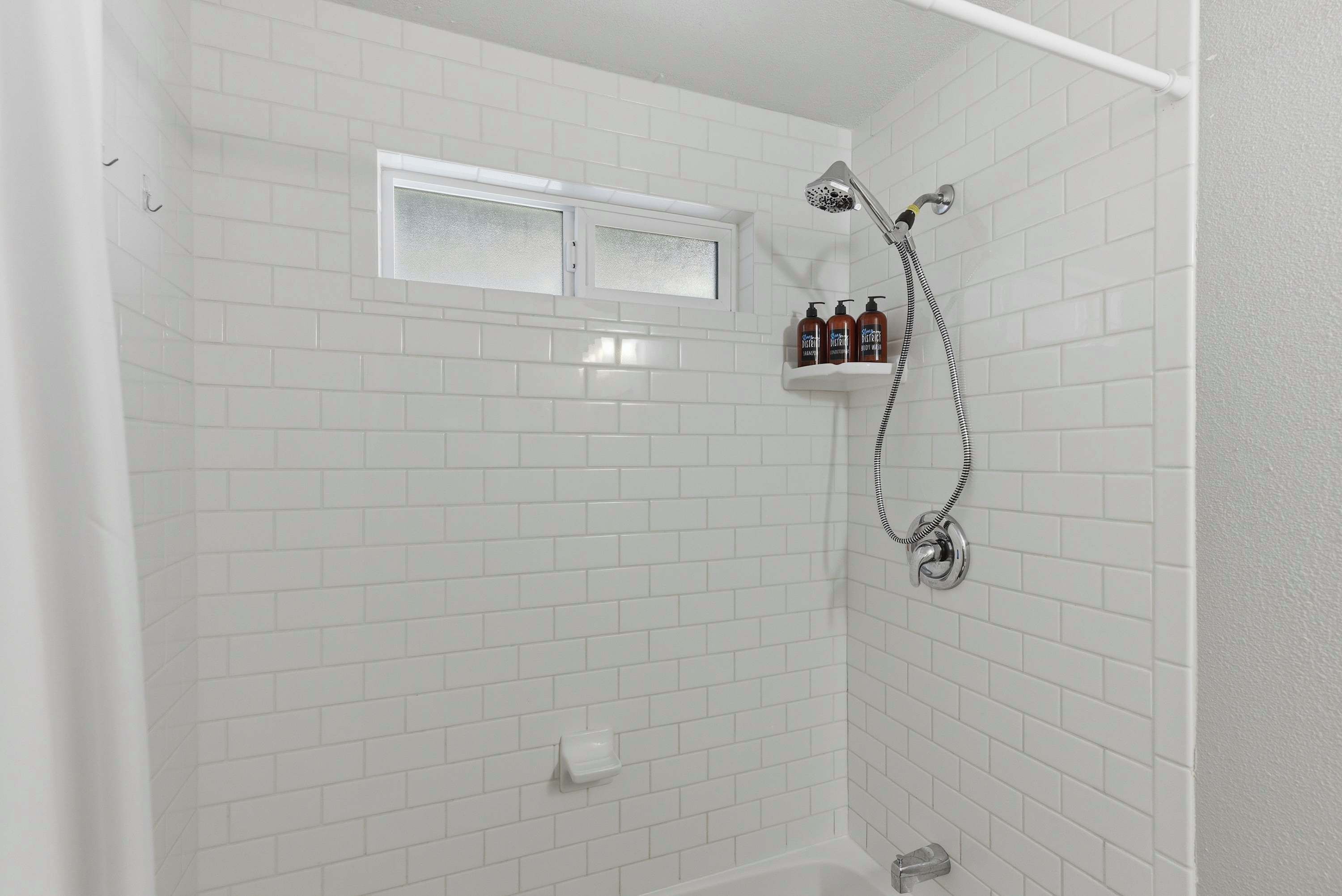 A shower area with white tiled walls, a window, and a shelf with toiletries.
