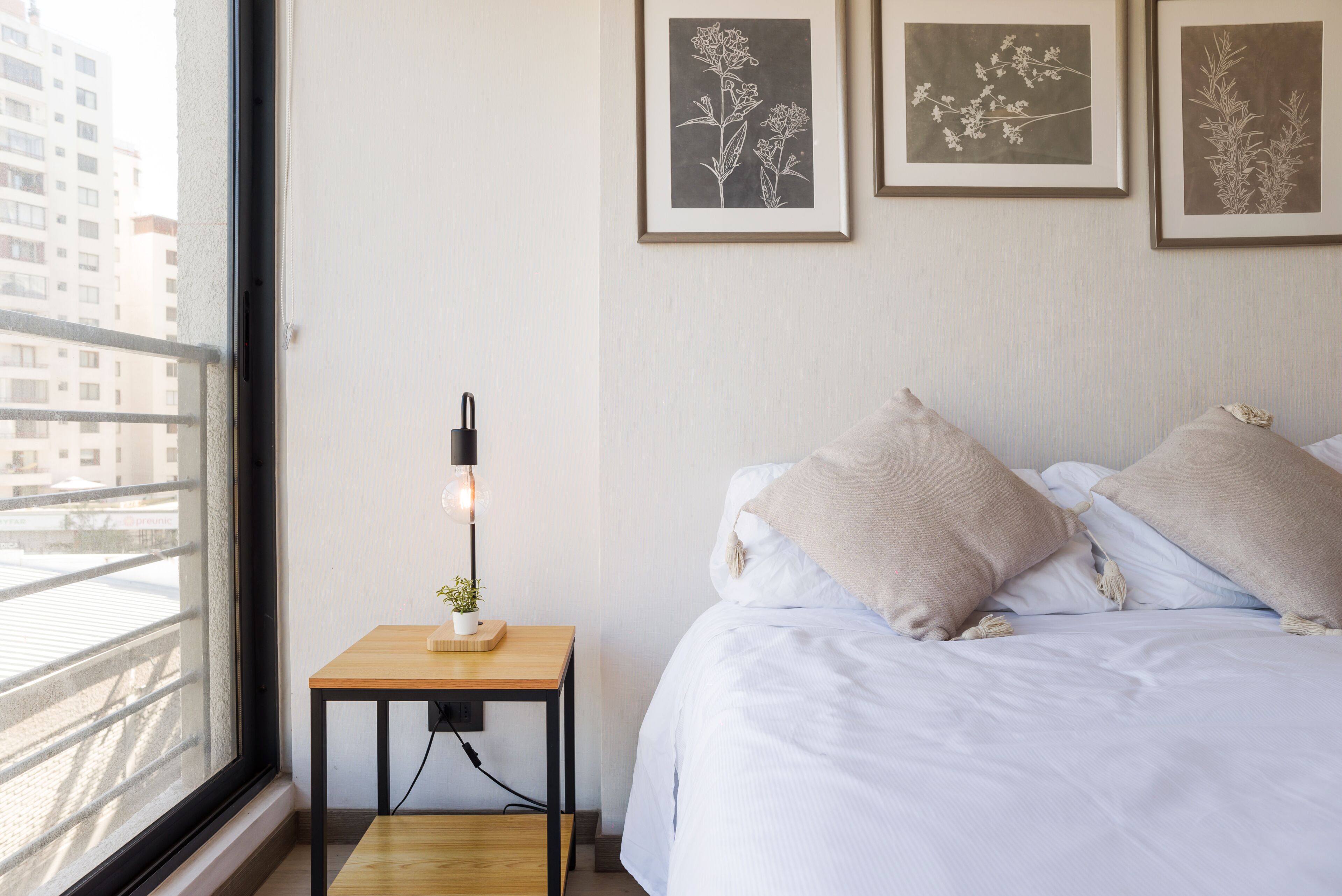 A bedroom with a bed, a bedside table with a lamp, and framed botanical prints on the wall.