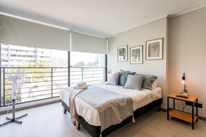 A modern bedroom with a large bed, a bedside table with a lamp, and a view of the city through a balcony. - Chic Modern Studio in Nunoa Vibran (Ñuñoa)