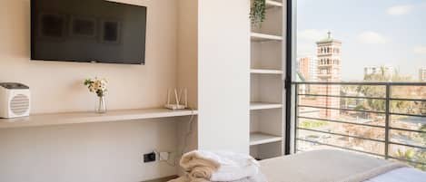 A modern bedroom with a large window, a flat-screen TV, a small plant, and a neatly made bed.