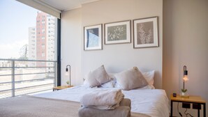 A hotel room with a large bed, a view of the city, and three framed pictures on the wall. - Chic Modern Studio in Nunoa Vibran (Ñuñoa)