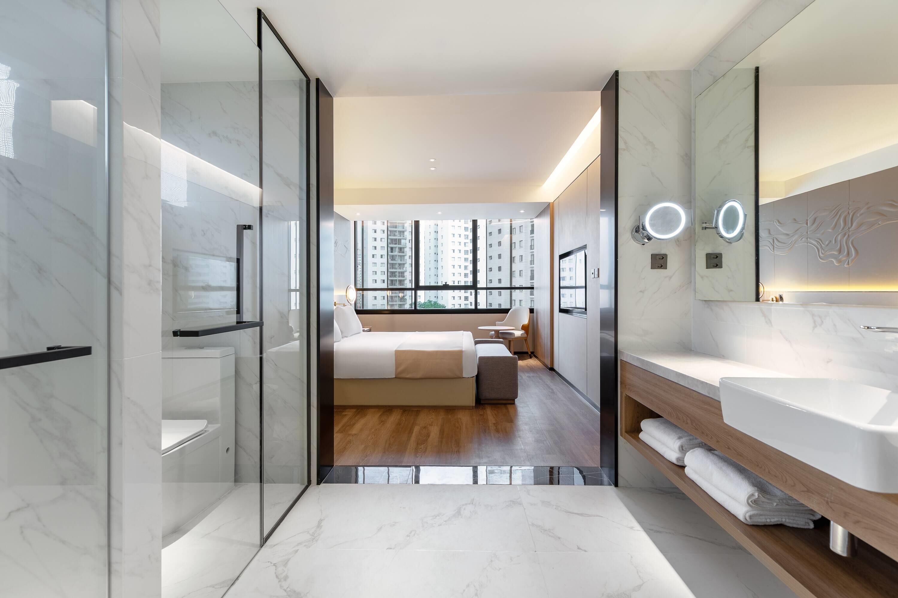Une chambre d’hôtel moderne dotée d’une douche en verre, d’un grand lit avec vue sur la ville, d’une coiffeuse en bois et d’un grand miroir avec éclairage intégré.