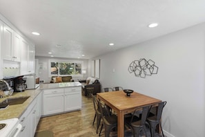 Cozinha moderna com armários brancos, área de jantar com mesa e cadeiras de madeira, e um objeto decorativo na parede.