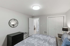 Quarto com cama, cômoda, espelho, porta e corredor que leva a outro espaço.