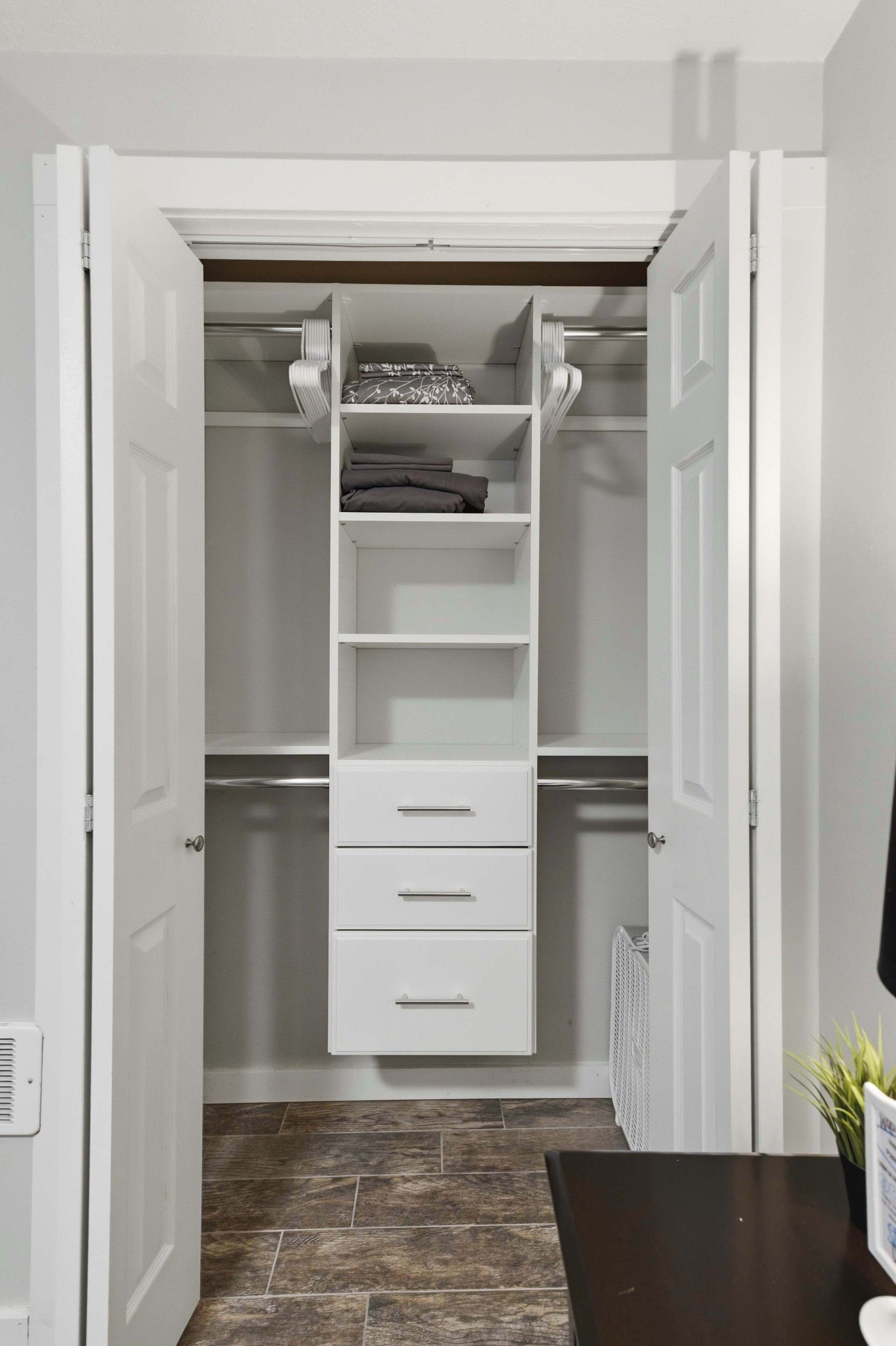 A walk-in closet with open doors, featuring a white shelving unit with folded clothes, a white radiator, and a dark wooden floor.