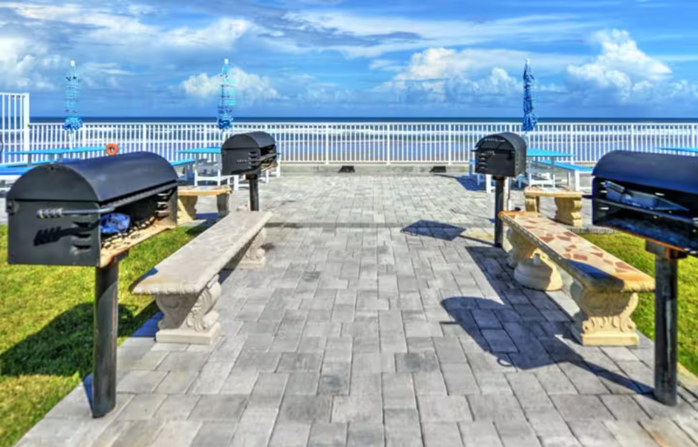 A paved outdoor area with barbecue grills and benches, overlooking a body of water.