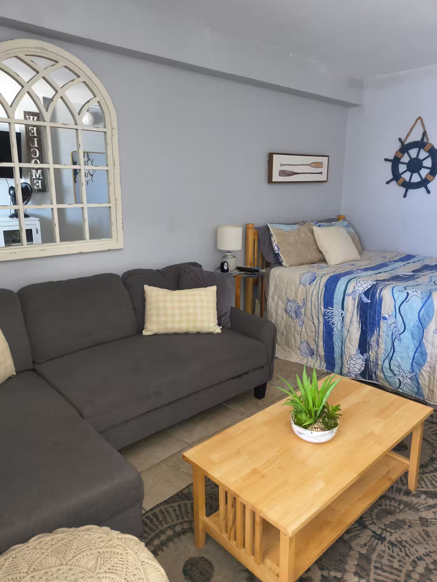 A living room with a sofa, a bed with a blue and white patterned bedspread, a wooden coffee table with a plant, and a large window with a decorative frame.