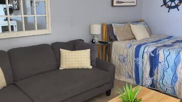 A living room with a sofa, a bed with a blue and white patterned bedspread, a wooden coffee table with a plant, and a large window with a decorative frame.