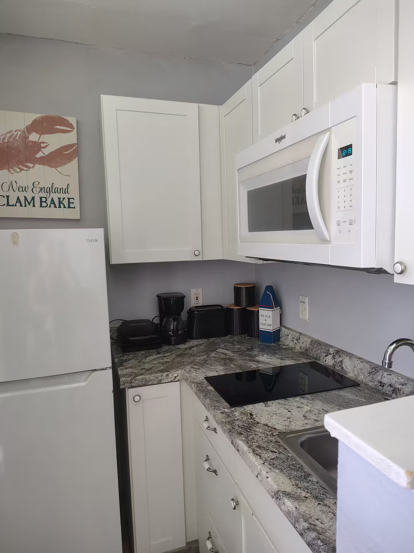 Cocina compacta con armarios blancos, microondas integrado, cafetera y lavabo.
