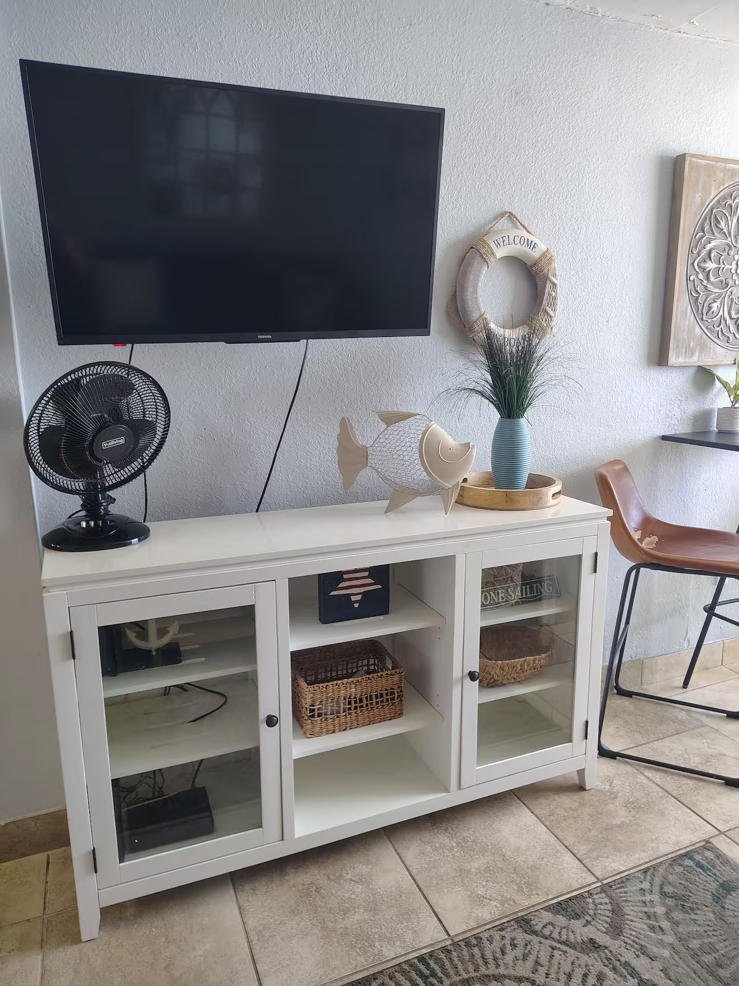 Sala de estar con televisor de pantalla plana sobre un mueble blanco con puertas de vidrio, un ventilador negro y objetos decorativos.