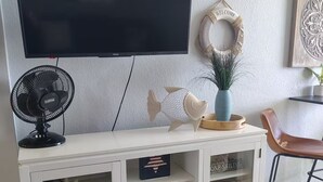 Sala de estar con televisor de pantalla plana sobre un mueble blanco con puertas de vidrio, un ventilador negro y objetos decorativos.