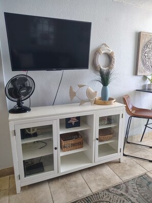 Sala de estar con televisor de pantalla plana sobre un mueble blanco con puertas de vidrio, un ventilador negro y objetos decorativos.