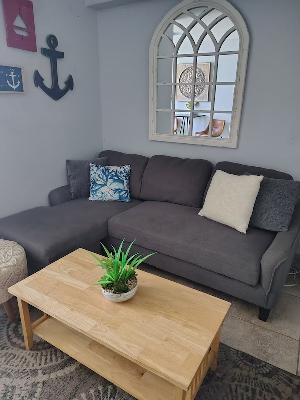 Una sala de estar con un sofá gris, una mesa de centro de madera con una planta y un ventanal decorativo con vista a un comedor.