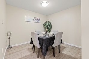 A dining area with a table set for four, surrounded by chairs, a plant, and a wall-mounted painting. - KING BED/ AC/ Henday /Whitemud (Edmonton)