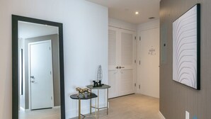 A modern hallway with a mirror, a small table with a decorative item, a wall-mounted artwork, and a door with a white shutter. - Cozy Apt - Corner Balcony w/360° Views (Hollywood)