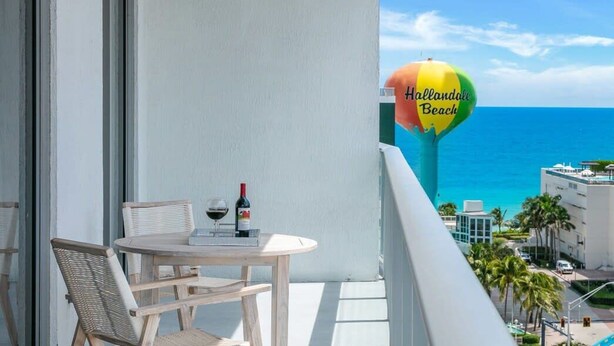 A balcony with a table and chairs, a bottle of wine, and a view of the ocean. - Cozy Apt - Corner Balcony w/360° Views (Hollywood)