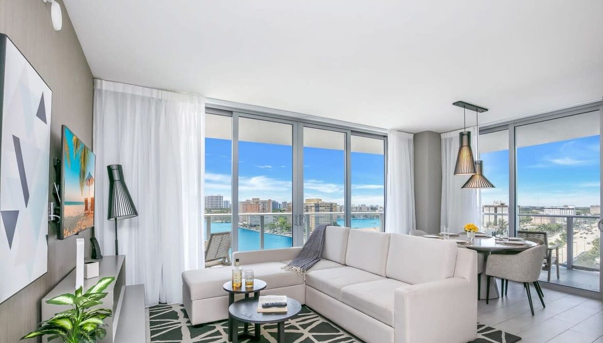 A modern living room with a sofa, coffee table, and dining area, featuring large windows with a city view.