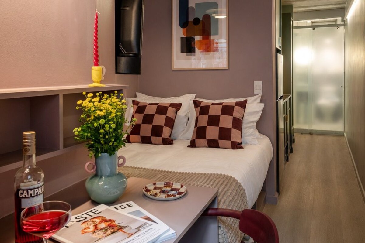 Ein kleines Hotelzimmer mit einem Bett, einem kleinen Tisch mit einer Zeitschrift, einer Flasche Alkohol und einer Vase mit Blumen.