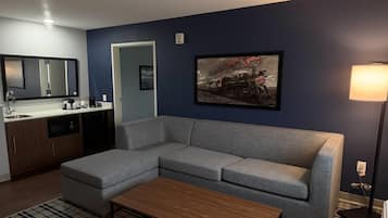 A modern living room with a grey sofa, a wooden coffee table, a kitchen area with a sink and coffee maker, and a framed picture on the wall.