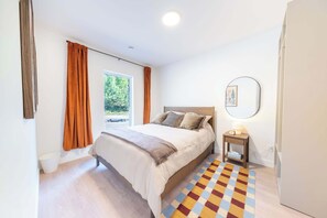 A bedroom with a bed, a bedside table with a lamp, a window with curtains, and a patterned rug.