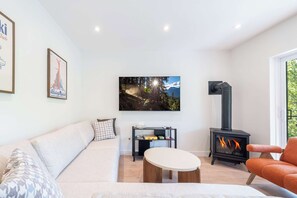 Ein modernes Wohnzimmer mit einer Couch, einem runden Couchtisch, einem an der Wand befestigten Fernseher, einem Kamin und einem Fenster mit Blick auf Bäume.