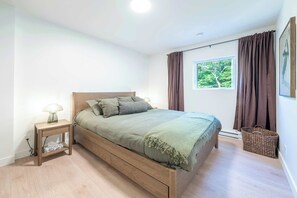 Ein Schlafzimmer mit einem hölzernen Bett, einem Nachttisch mit Lampe, einem Fenster mit Vorhängen und einem Wandbild.