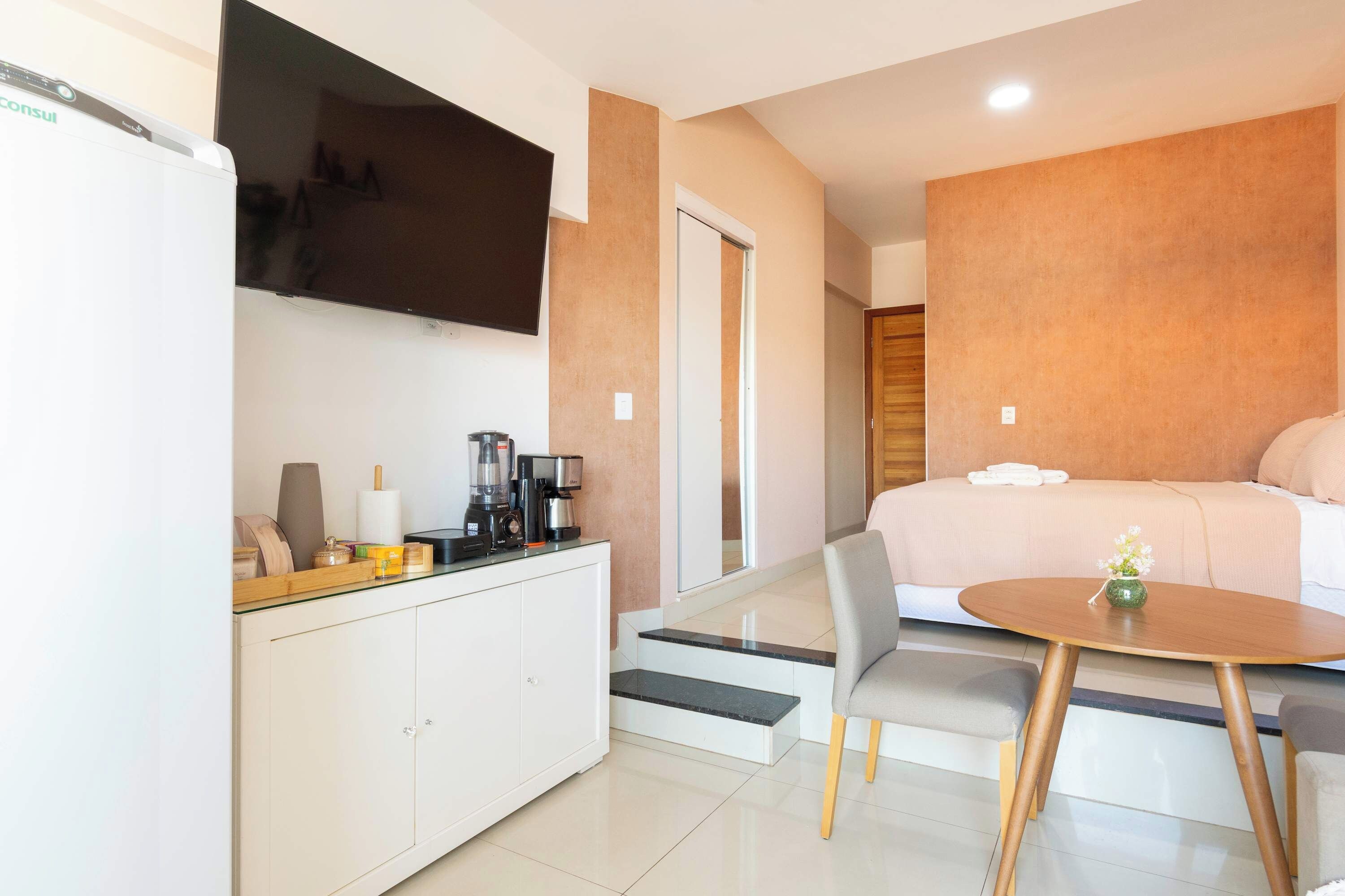 A modern hotel room with a bed, a small dining table, a TV, and a kitchenette.