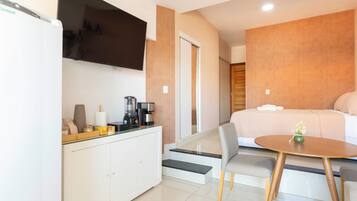 A modern hotel room with a bed, a small dining table, a TV, and a kitchenette.