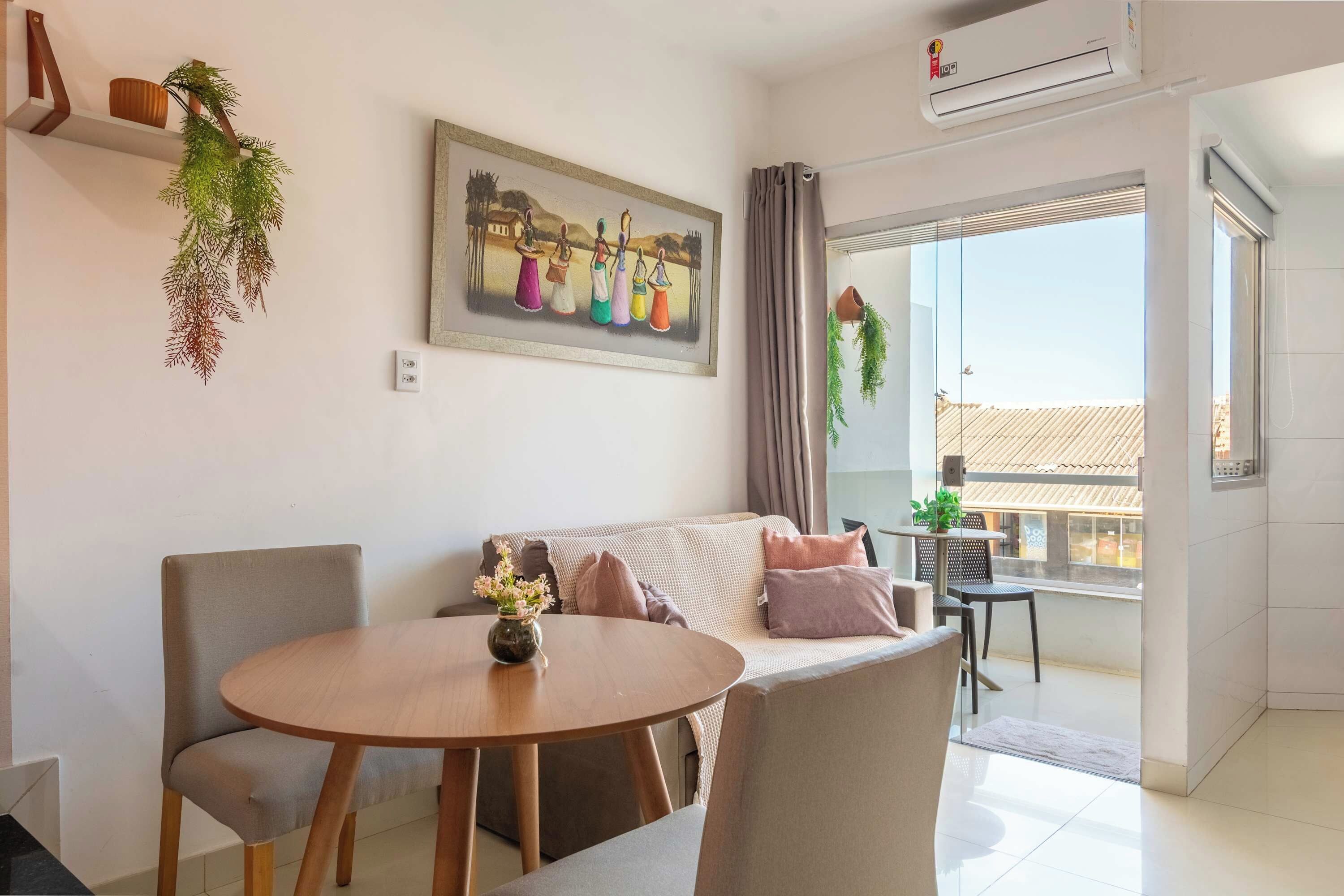 A modern living room with a round wooden table, a sofa with cushions, a painting on the wall, and a balcony with a view.