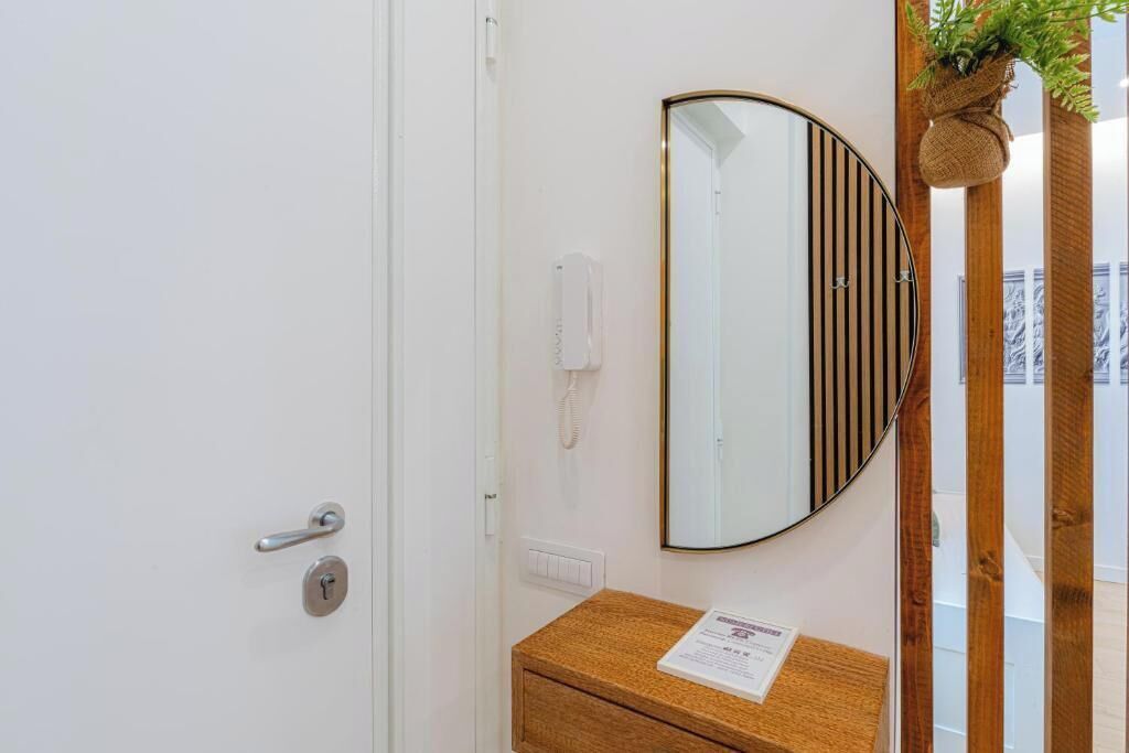 A hotel room with a wooden door, a mirror with vertical stripes, a wooden bedside table, and a plant hanging on the wall.