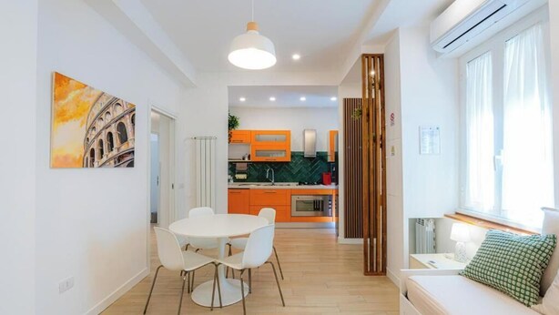 A modern kitchen with orange cabinets, a dining area with a white table and chairs, and a living area with a sofa and a window. - Contessa Elegance Inn Vacation Rental, Rome (Roma)