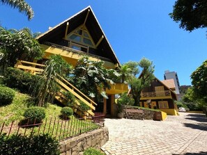 A yellow building with a balcony, surrounded by greenery and a paved area.