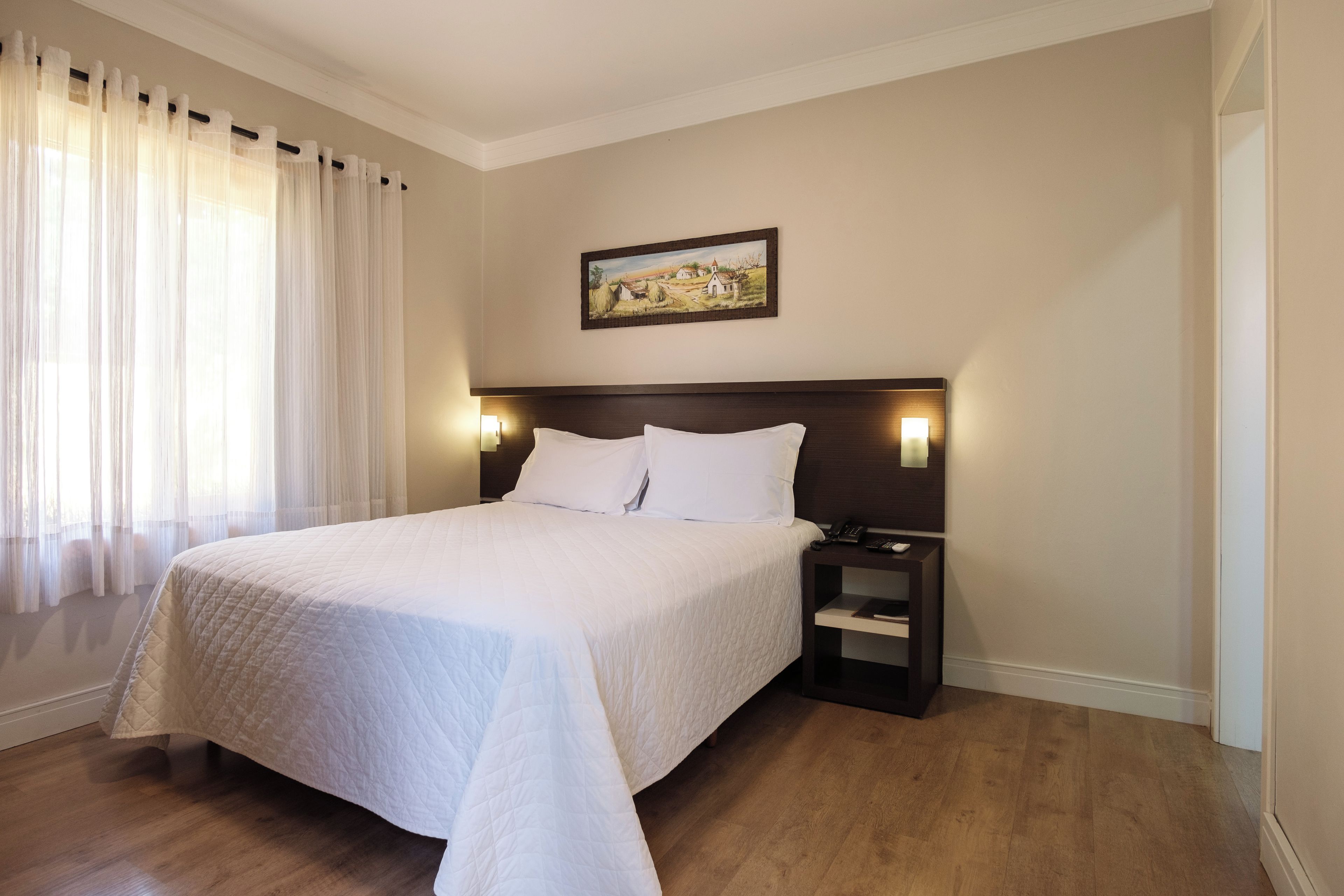 A hotel room with a large bed, a nightstand, a painting on the wall, and a window with curtains.