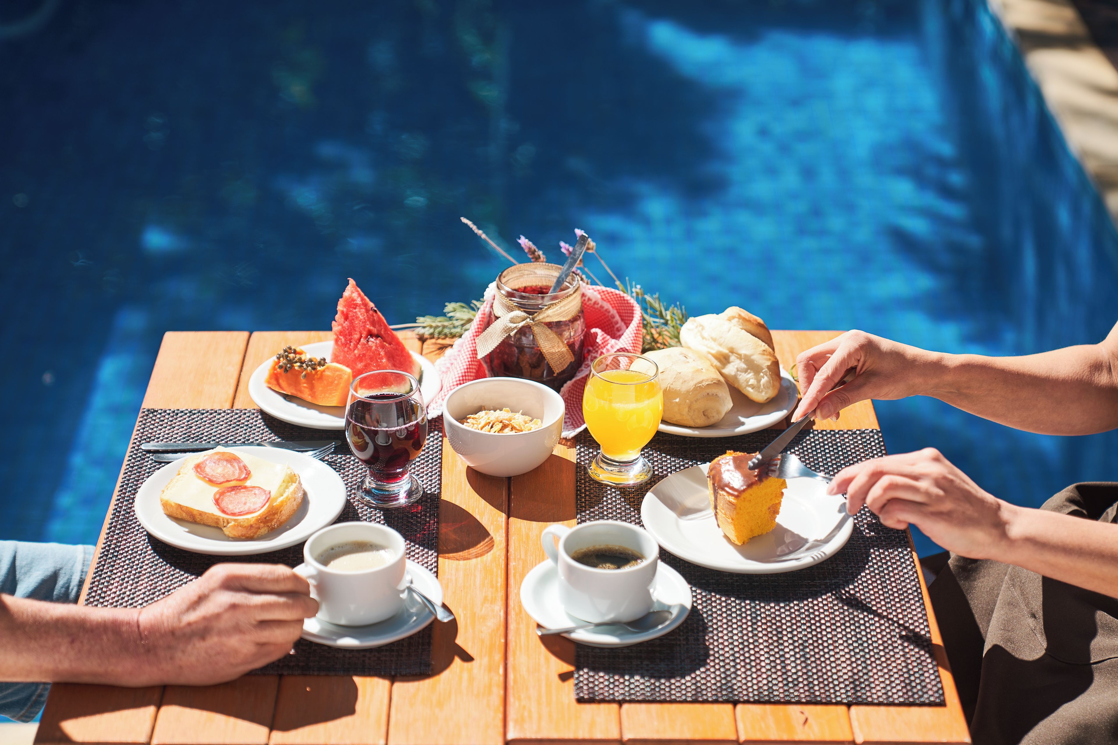 Una mesa de comedor con alimentos para el desayuno, una copa de vino tinto y un vaso de jugo de naranja junto a una piscina.