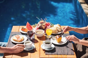 A dining table with breakfast items, a glass of red wine, and a glass of orange juice by a poolside.