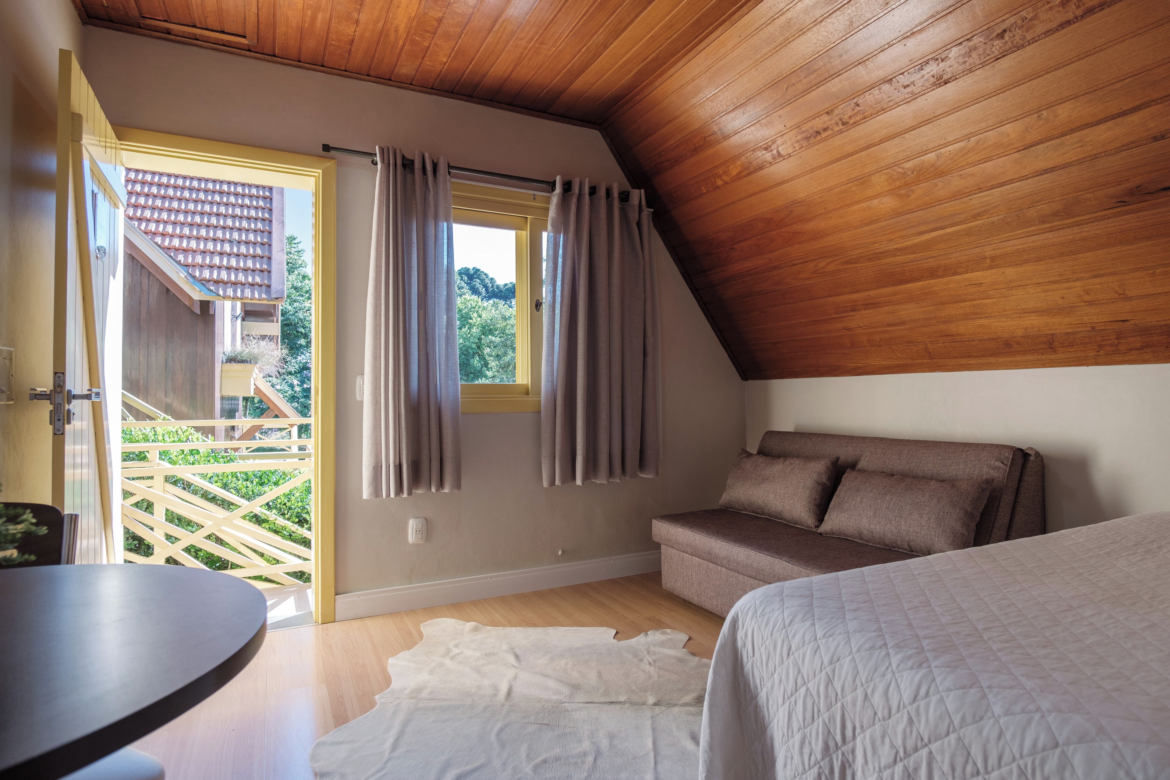A bedroom with a wooden ceiling, a sofa, a bed, and a view of a garden through a window.