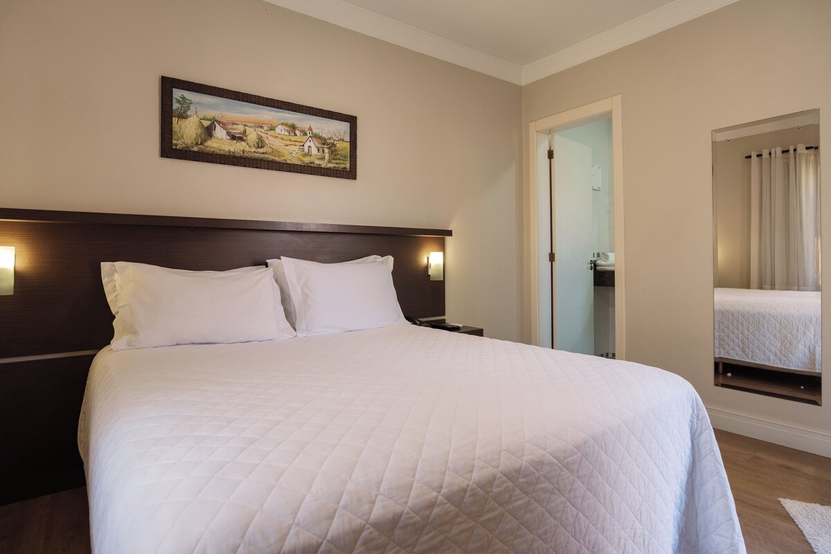 A hotel room with a large bed, a nightstand, a painting on the wall, and a mirror reflecting the bed.