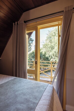 Un dormitorio con cama, ventana con cortinas y vistas a un balcón y árboles.