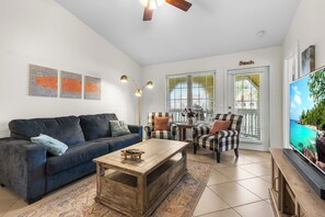 Un salon comprenant un canapé, des fauteuils, une table basse et un téléviseur. Il y a trois lampes fixées au mur et un ventilateur de plafond.