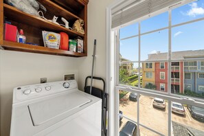 A laundry area with a washing machine, a shelf with cleaning supplies, and a view of a parking lot through a window. - Salt & Summer at the Dawn condos with Ocean and Pool Views!  Family friendly! (Galveston)